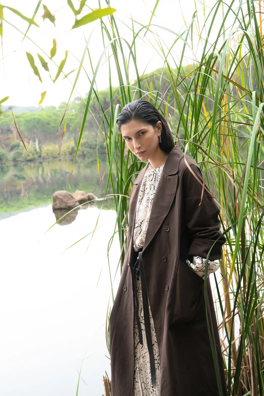 Bruma: la modelo entre los juncos del embalse, con abrigo marrón largo sobre vestido estampado