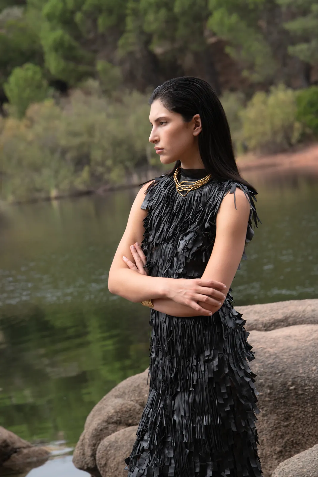 Bruma: vestido negro de flecos y collares dorados, de brazos cruzados junto al agua